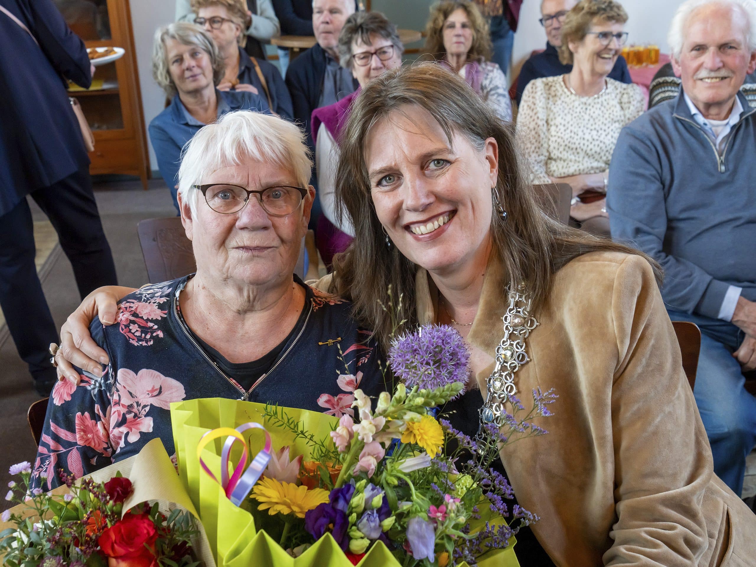 Twee vrouwen kijken lachend naar de camera. De mevrouw links heeft een bos bloemen in haar handen. Op de achtergrond zit een groep mensen die lachen en blij kijken.