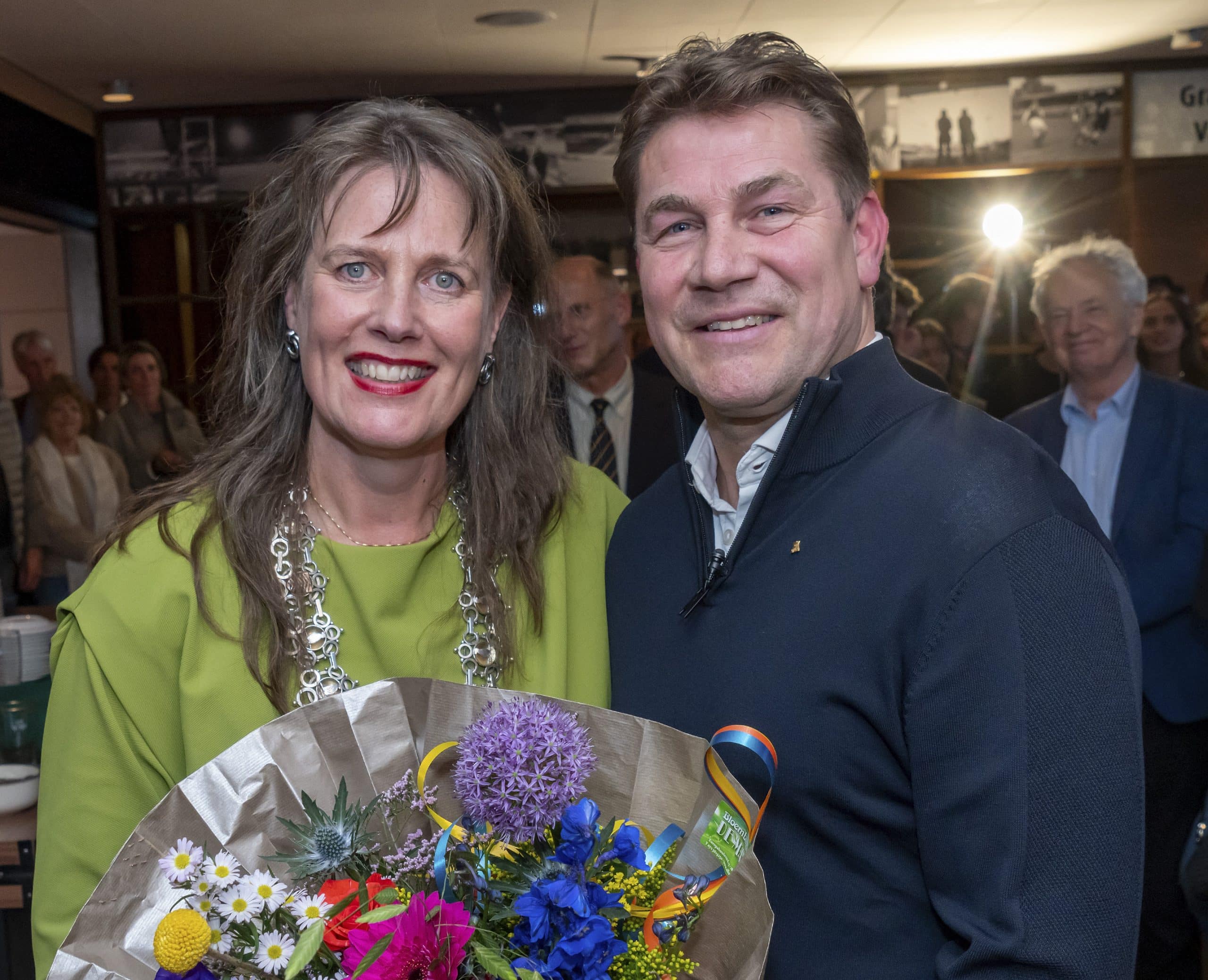 Burgemeester Anja Schouten en Max Huiberts, voormalig technisch directeur van AZ, kijken in de camera. Op de achtergrond een groep mensen.