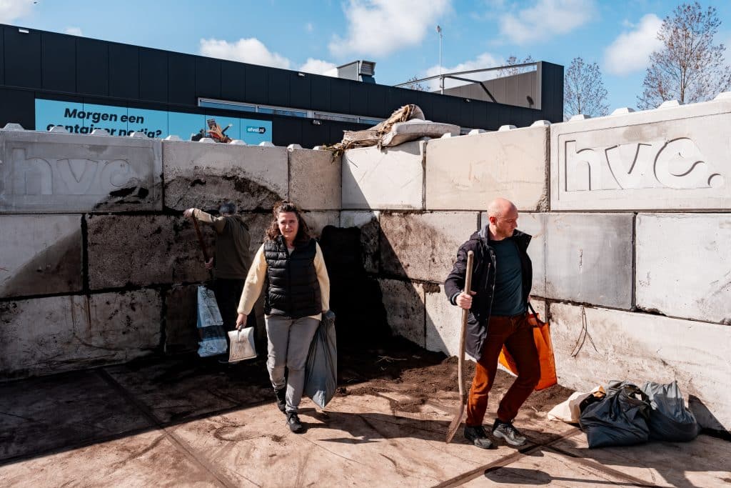 2 mensen lopen weg van een berg compost met een schep en volle zakken compost in hun handen.