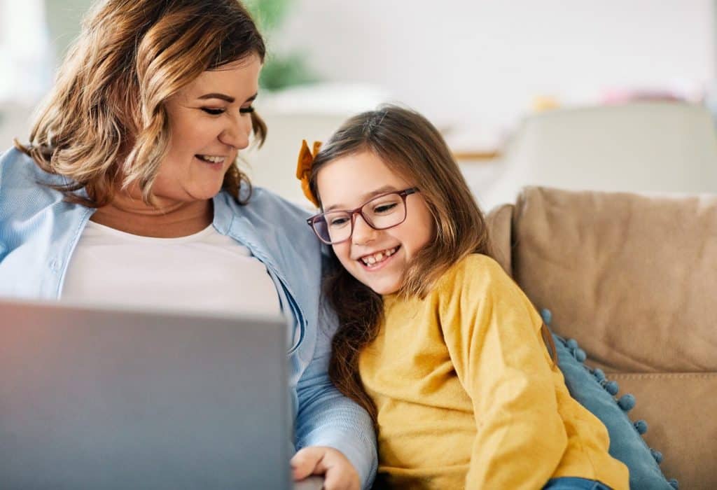 Een vrouw zit met laptop op schoot naast haar dochter op de bank. De moeder lacht naar haar dochter en de dochter kijkt lachend naar de laptop.