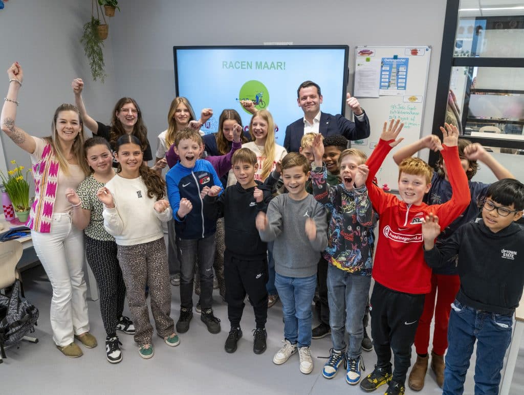Wethouder Christiaan Peetoom samen met leraren, begeleiders en kinderen in een klaslokaal voor een scherm waarop staat Racen maar!