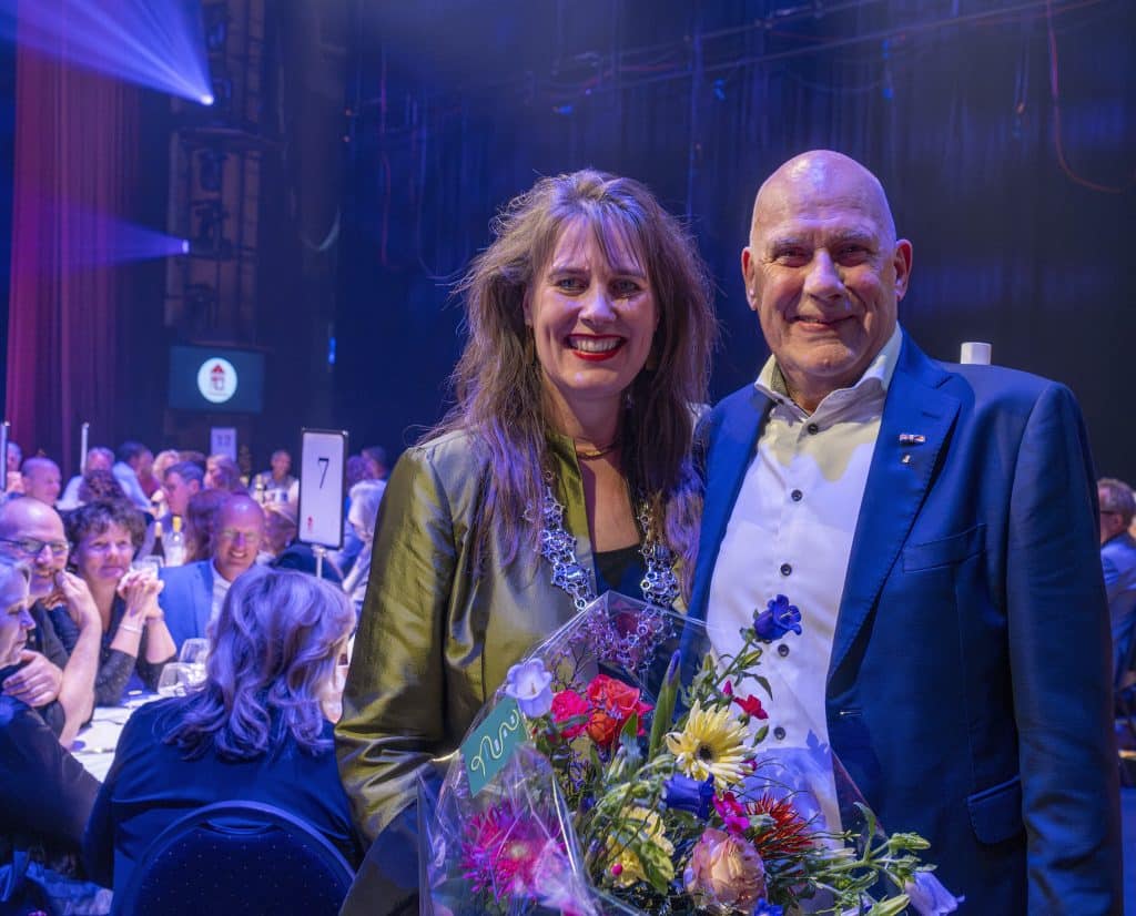 Burgemeester Anja Schouten staat naast Jaap Hoogland tijdens het Praethuysgala met een bos bloemen en op de achtergrond mensen die aan tafel zitten