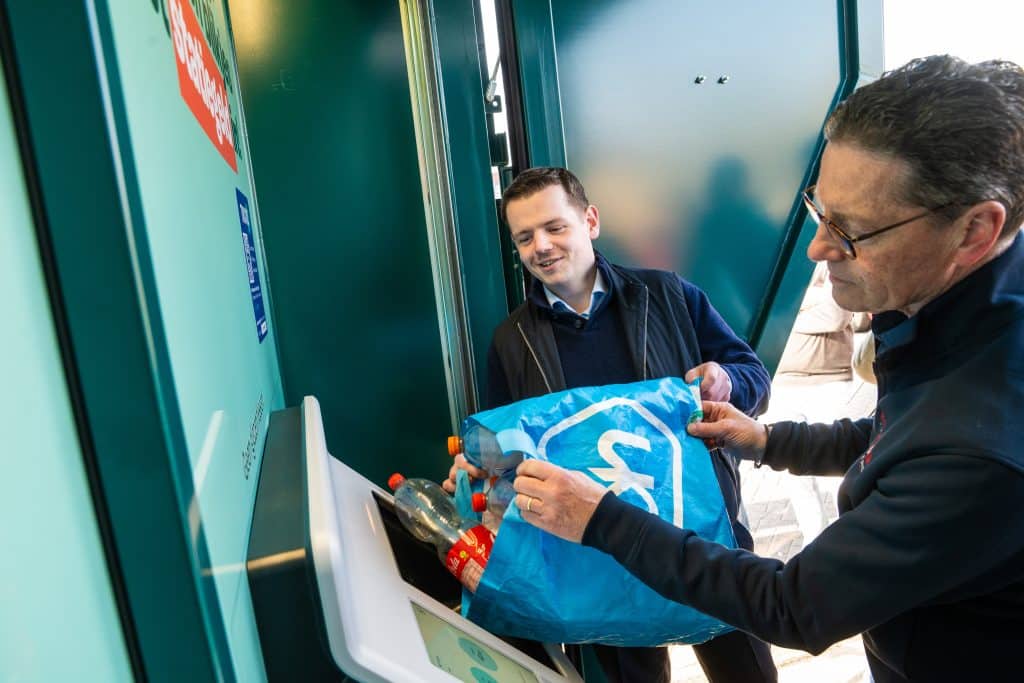 Wethouder Christiaan Peetoom en Hans de Boer gooien samen een tas vol flessen en blikjes in de opening van de statiegeld bulkmachine