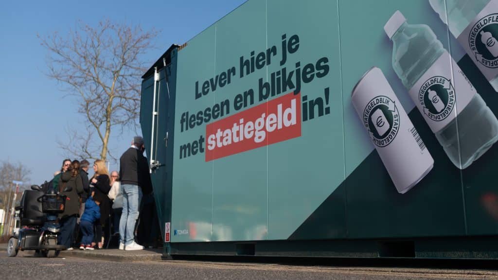 Grote groene statiegeld bulkmachine in de vorm van een zeecontainer met daarop de tekst Leveri hier je flessen en blikjes met statiegeld in! Links voor de machine staan mensen. 