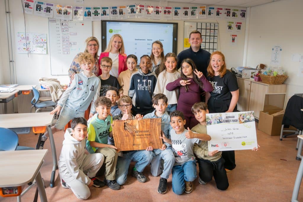 Klas van school Jules Verne in hun klaslokaal als groep bij elkaar met docenten en wethouder Christiaan Peetoom. Ze houden hun prijs vast: een cheque en een plakkaat.