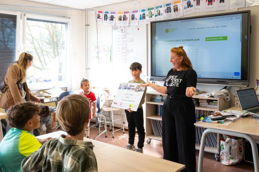 Docent van Race against waste voor de klas met daarnaast een leerling die een grote cheque vasthoudt. Op de voorgrond kinderen aan tafeltjes. 