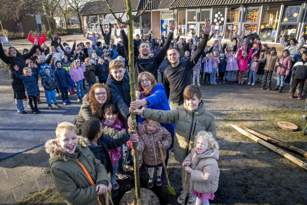 Foto van groep volwassenen en kinderen die een boom planten op het schoolplein van een basisschool