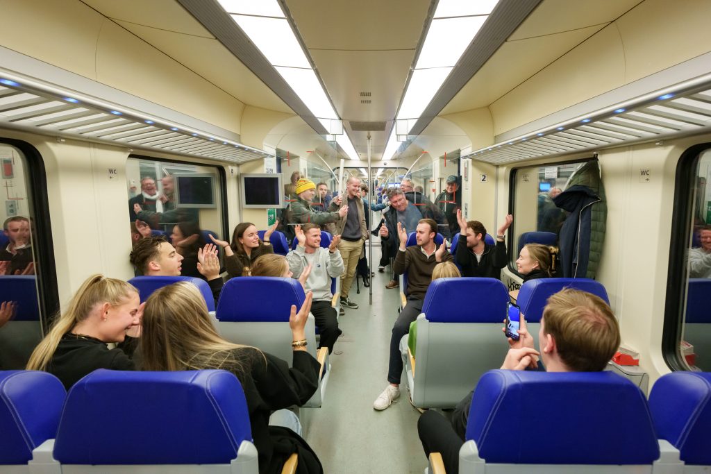 Een groep reizigers zit en staat in een trein en applaudisseert.