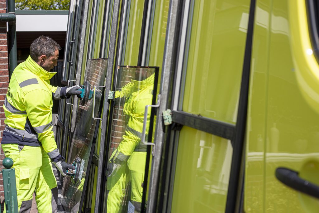 Man pakt glasplaat van zijn bedrijfsauto. Hij draagt beschermende kleding en handschoenen.