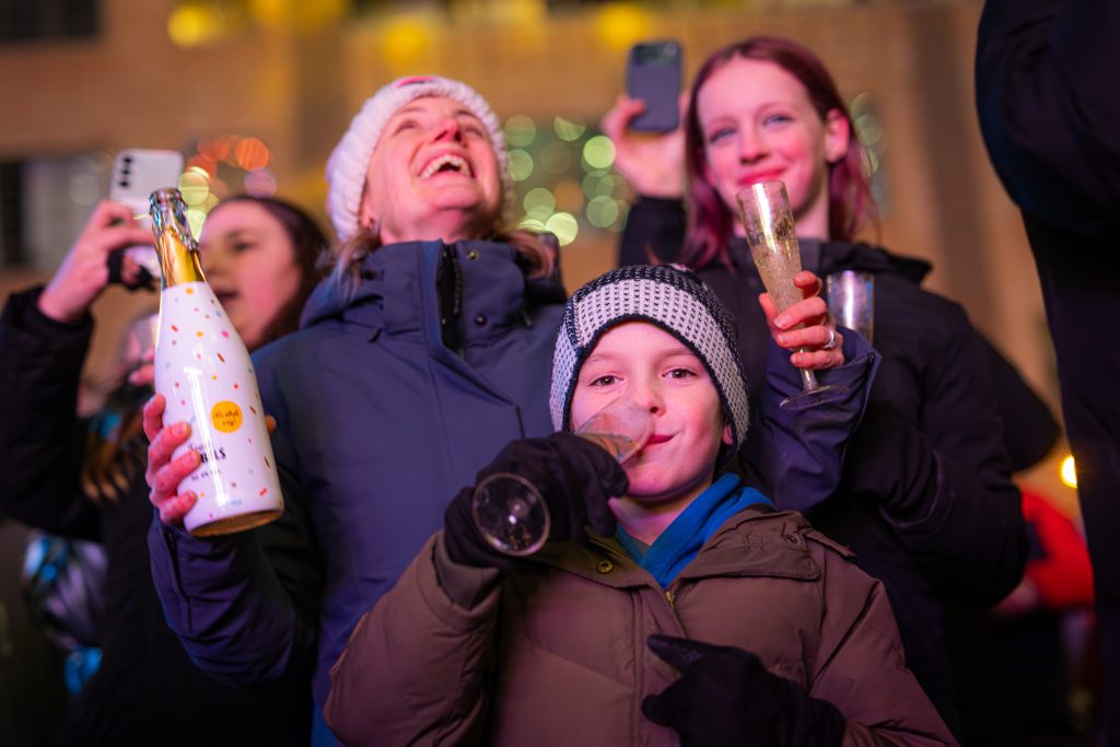 Een lachende groep feestvierders, onder wie een kind met een wintermuts, viert buiten de jaarwisseling.