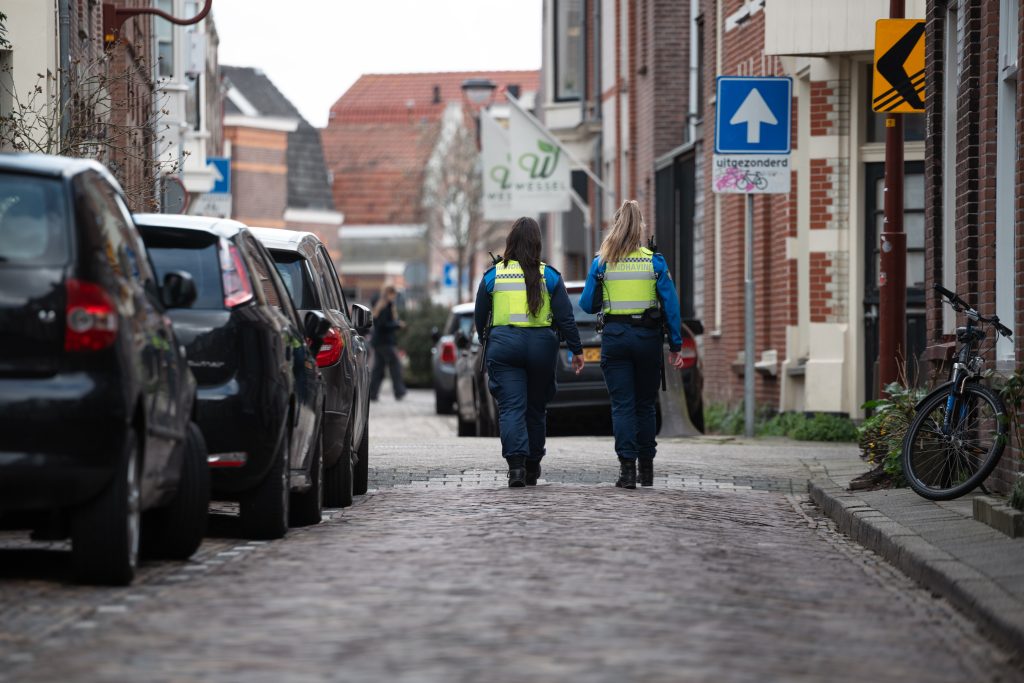 Twee handhavers lopen naast elkaar door een woonstraat met geparkeerde auto's en bakstenen huizen.