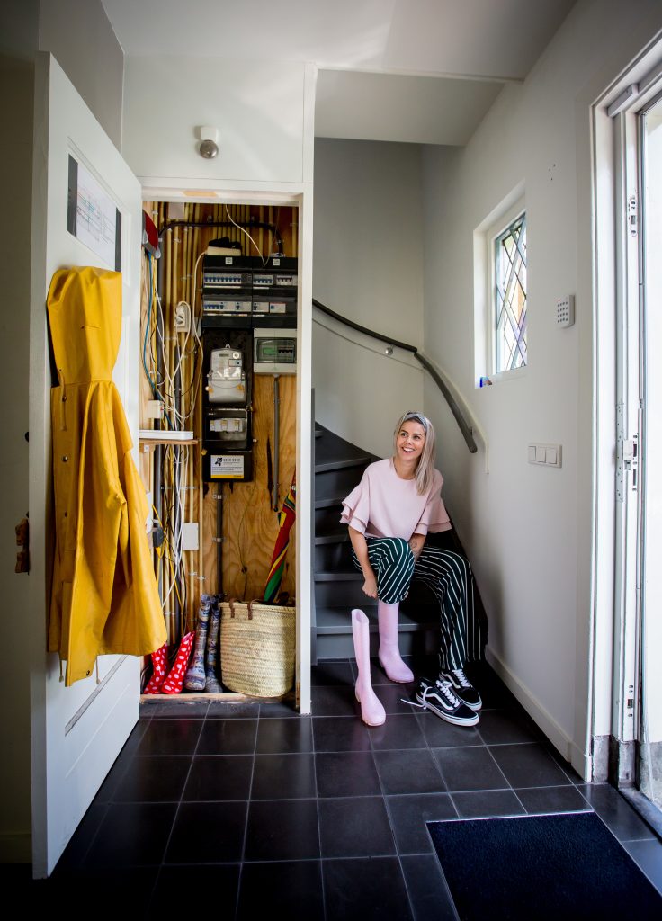 Vrouw zit onder aan trap in hal van huis en trekt een regenlaars aan. Links in beeld een meterkast met de deur open en op de voorgrond hangt een regenjas aan de kapstok.