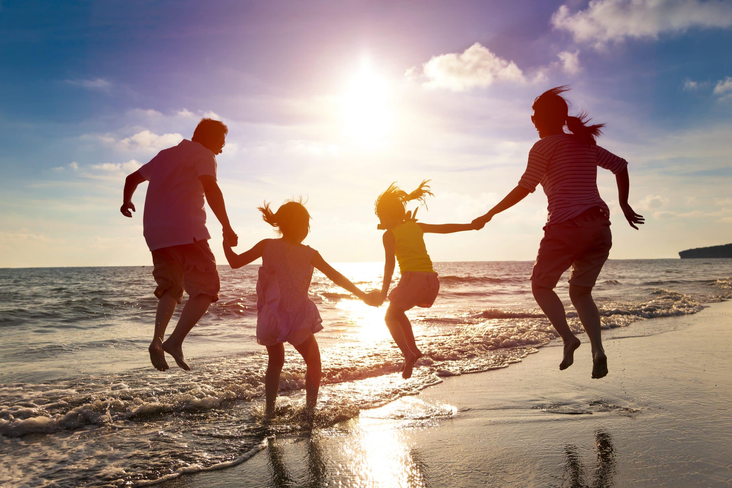 Twee volwassenen en twee kinderen houden elkaars hand vast terwijl ze in de lucht springen in de branding, terwijl de zon ondergaat.