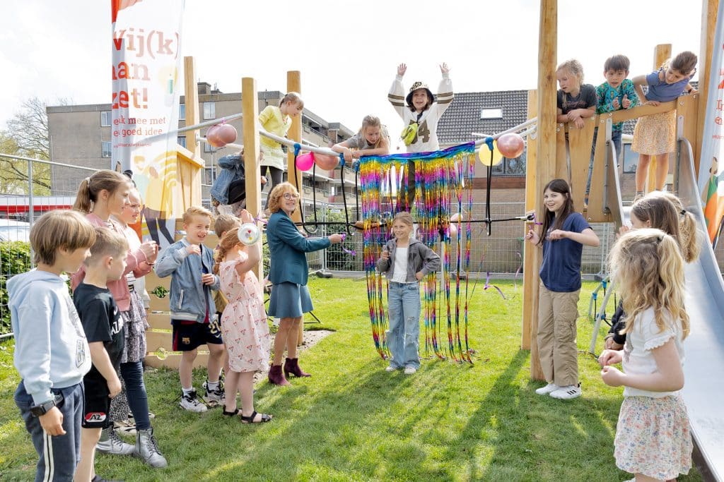 Er staan meerdere kinderen op, rond en naast het nieuwe houten speeltoestel. Het speeltoestel heeft een glijbaan en een touwbrug.