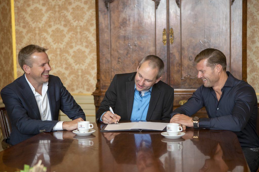 Wethouder Gijsbrecht zit aan een tafel en ondertekent een papier, aan beide zijdes zitten twee mannen lachend toe te kijken.