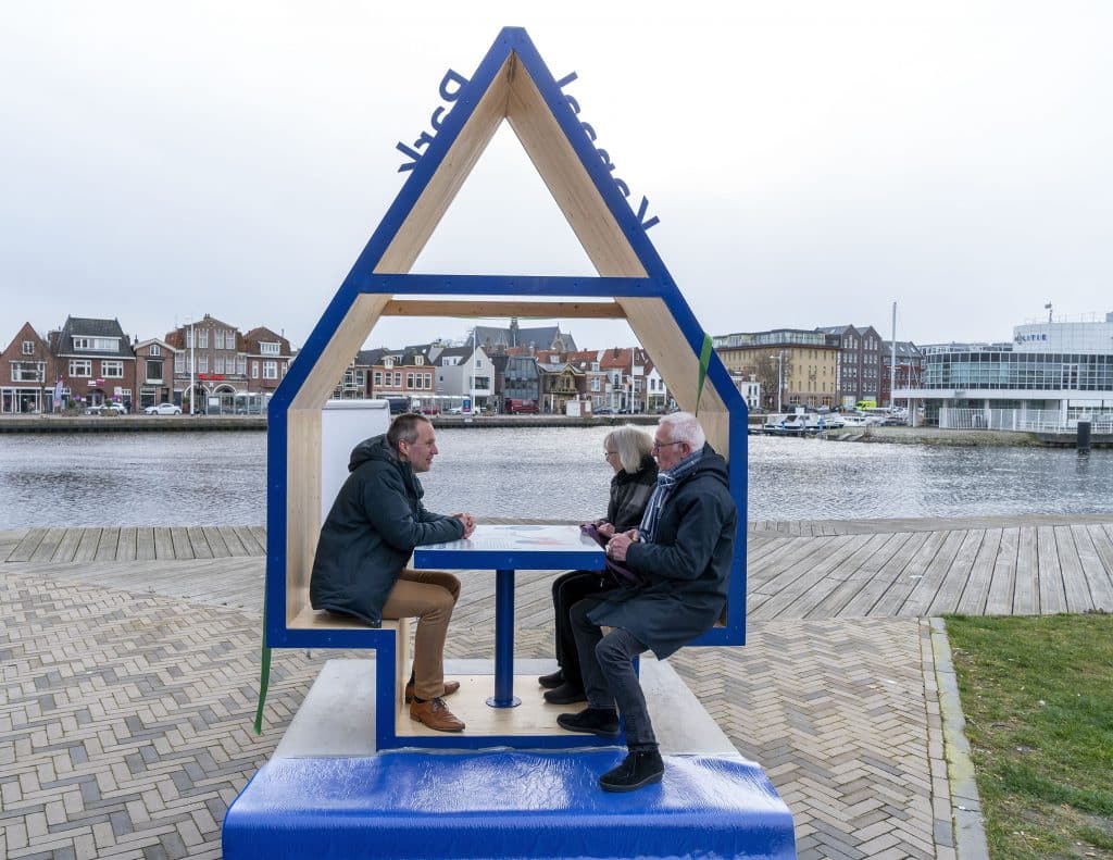 Wethouder Gijsbert van Iterson in gesprek met bewoners over hun wensen voor Alkmaars Kanaalpark. Ze zitten in een huisvorming hokje aan een tafel langs het kanaal.