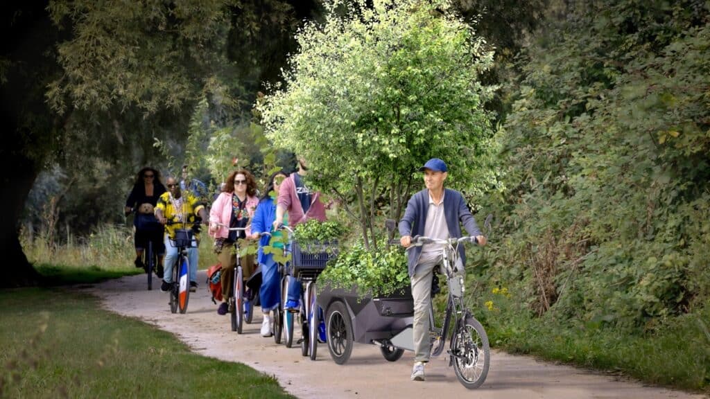 Een man, Bruno Doedens  staat stil met zijn fiets met een fietskar met een flinke boom erin. Achter hem fietsers die hem volgen. Ze staan op een fietspad in een groene omgeving. 