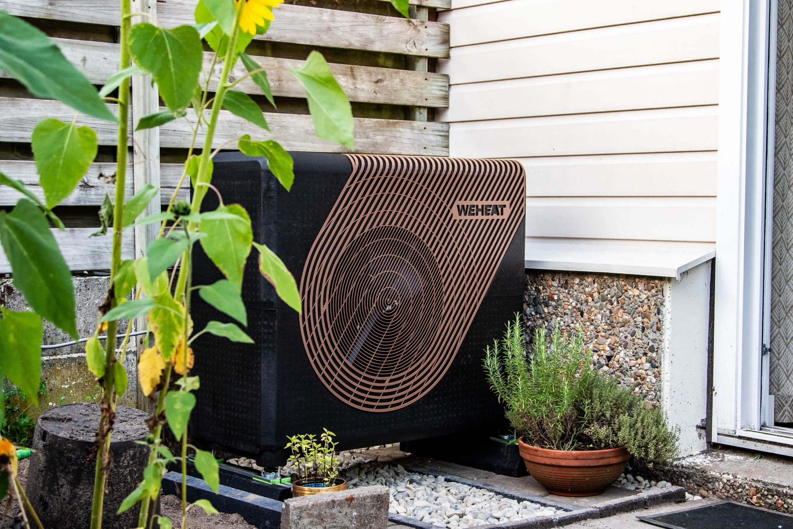 Een buitenunit van een warmtepomp die staat in de achtertuin van een woning met eromheen enkele planten.
