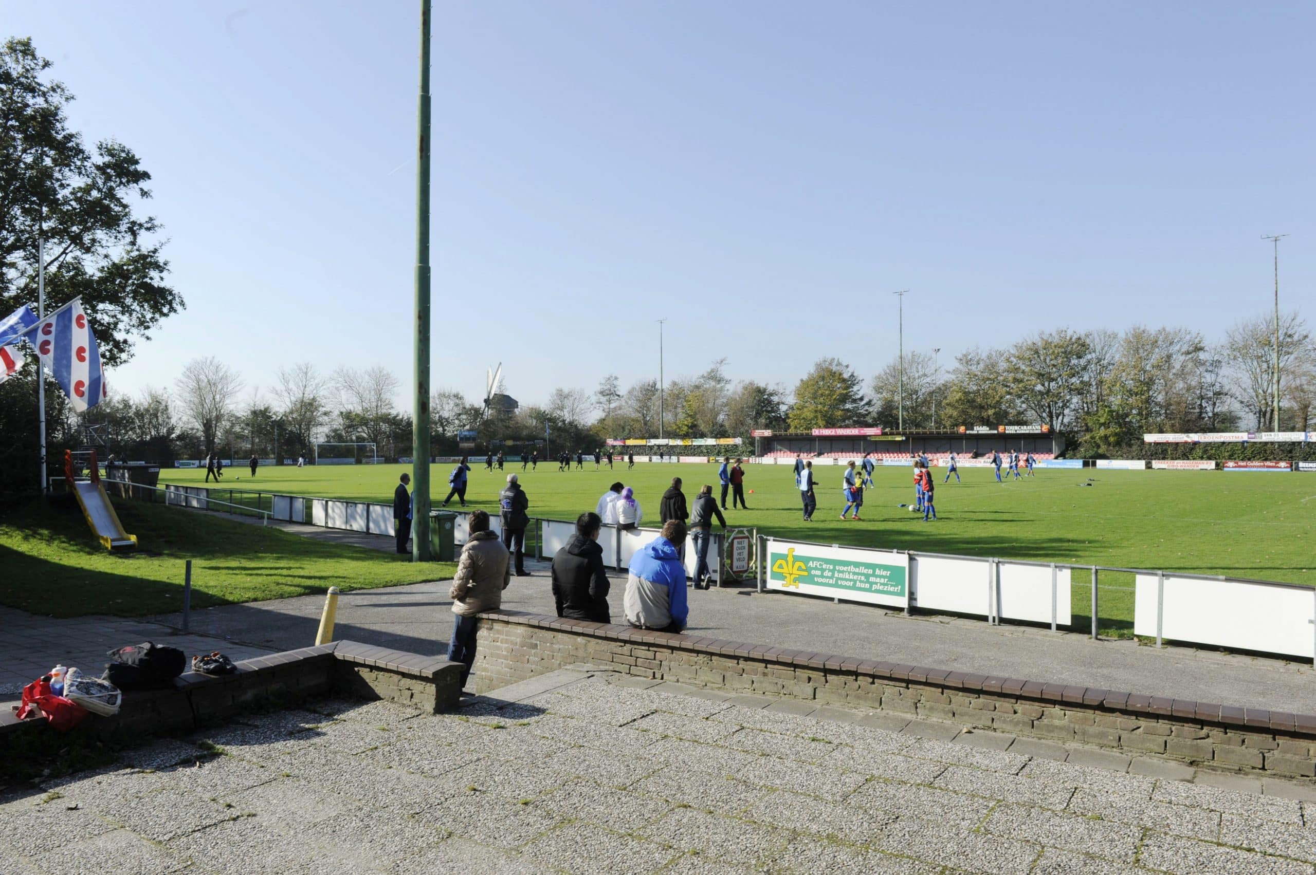 Voetbalveld met op de voorgrond een aantal mensen die kijken naar een voetbaltraining