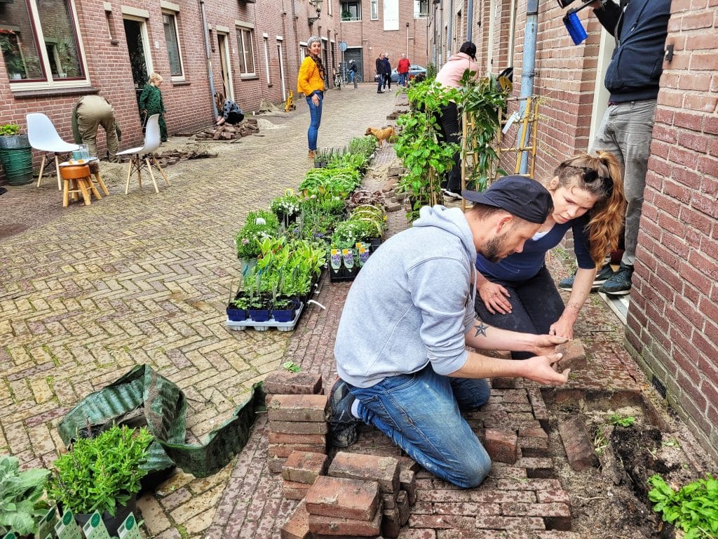 Straat met woonhuizen, mensen halen stenen uit de straat en zetten daar planten in de grond.