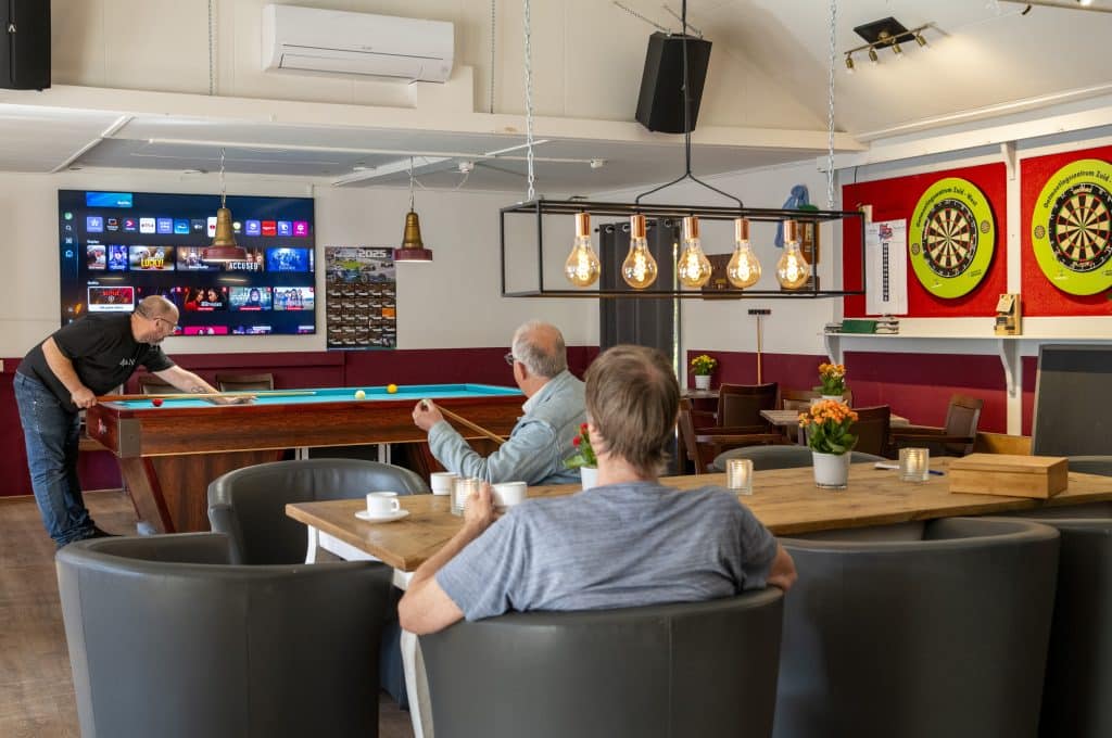 Café-achtige setting met biljart, man is aan het biljarten, twee kijken toe. 