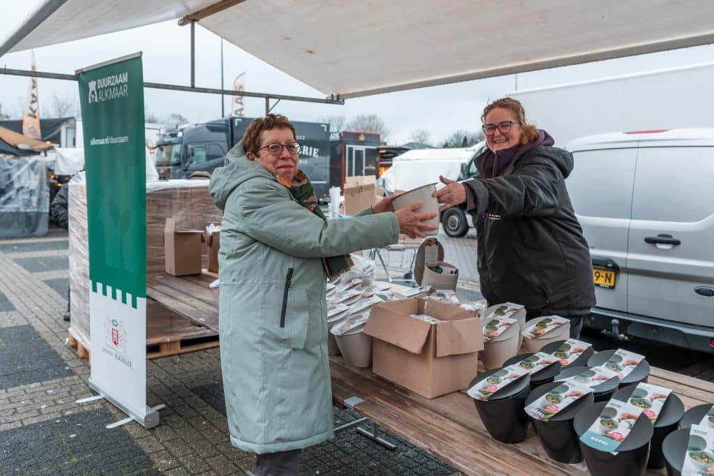 Afvalcoach geeft vanaf de marktkraam een gft-bakje aan een inwoner. Op de kraam staan witte en zwarte gft-bakjes. Naast de kraam een banner van Duurzaam Alkmaar.