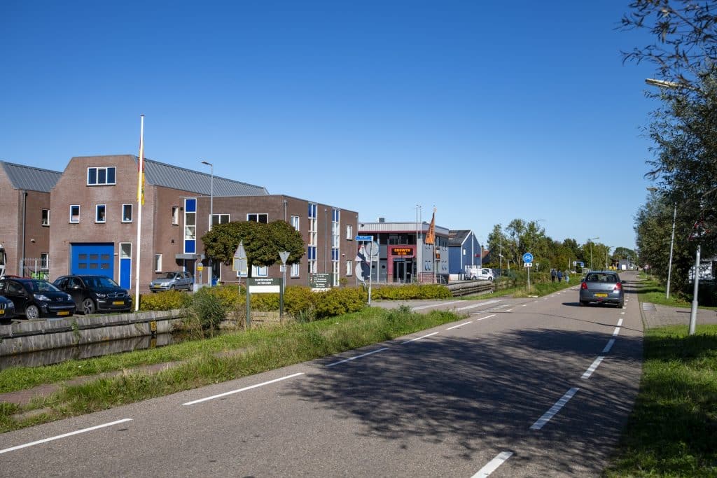 Op de voorgrond een asfaltweg met een auto die van je wegrijdt. Links naast de weg gebouwen van bedrijventerrein de Volger in de Rijp