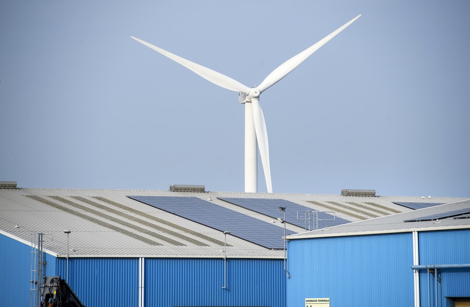 Op de voorgrond een grote bedrijfshal met zonnepanelen op het dak, op de achtergrond een grote windmolen. 