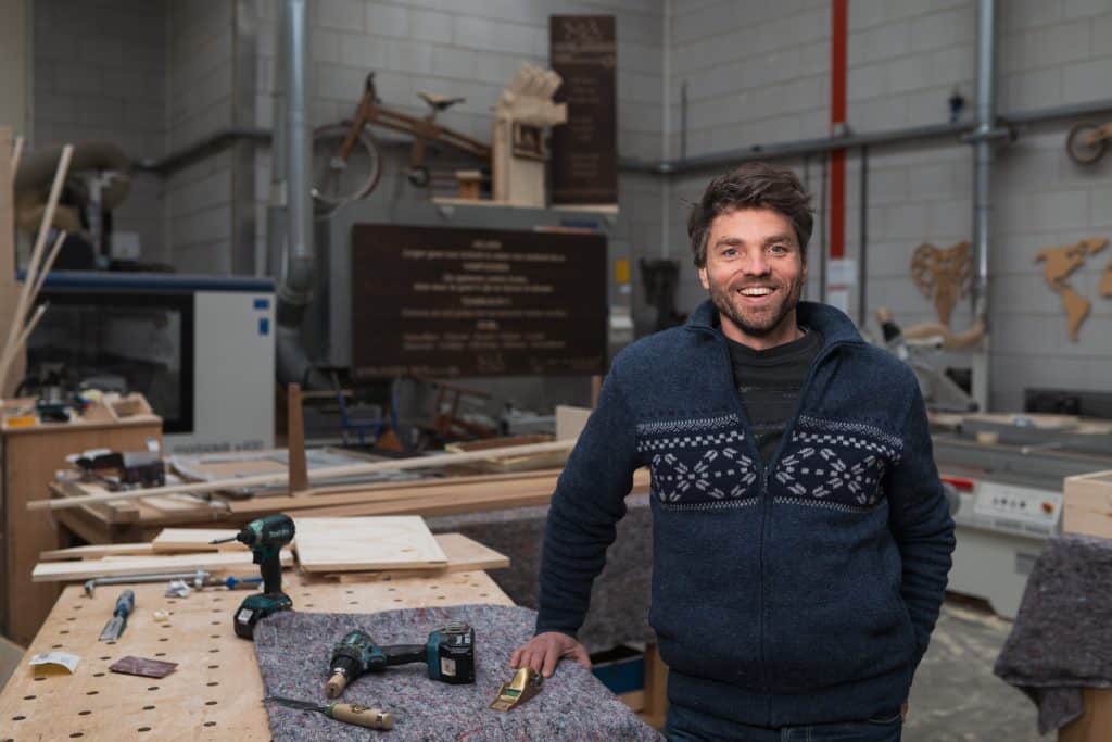 Jan Gunneweg in zijn werkplaats. Hij staat naast een werkbank met daarop gereedschap en hout. 