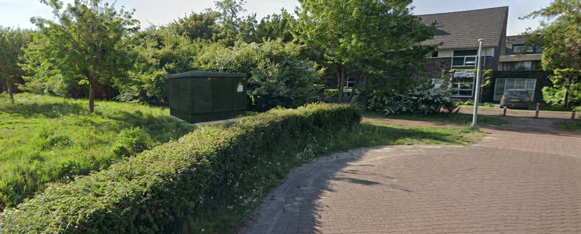 Voorbeeld elektriciteitshuisje Branding. Een beetje verscholen achter bosjes. Donkergroen en rechthoekig. Omringd met bosjes en bomen.