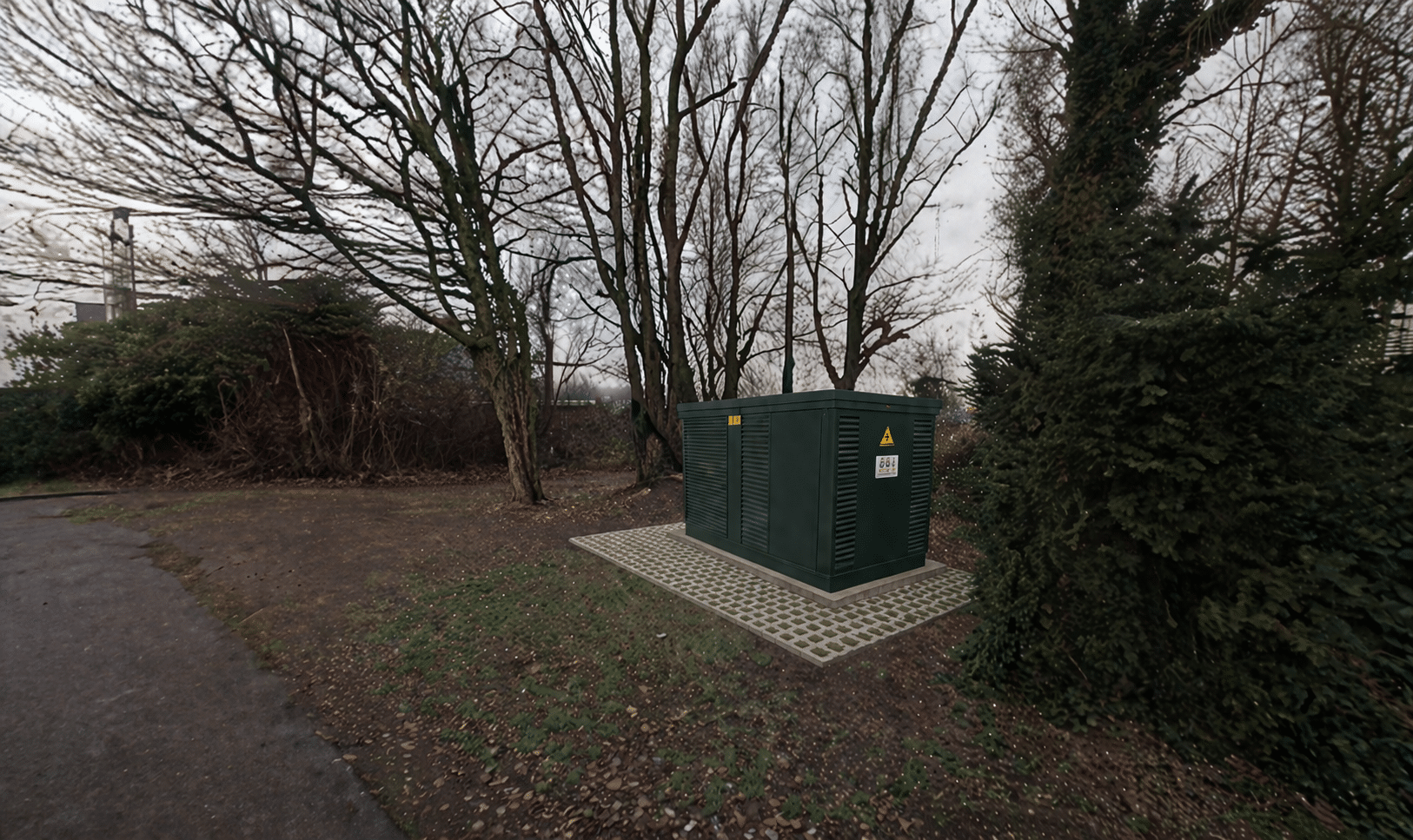 Voorbeeld elektriciteitshuisje Zeglis. Naast een pad.  Donkergroen en rechthoekig. Omringd met bosjes en bomen.